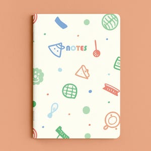 Pode incluir: Um caderno de cor creme com um padrão repetitivo de pequenos ícones coloridos desenhados à mão, incluindo círculos, alimentos e utensílios de cozinha. A palavra "NOTES" está impressa no centro em letras multicoloridas arredondadas.