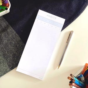 Op de afbeelding: Een wit rechthoekig notitieblok voor het bijhouden van gewoontes met het woord HABITS bovenaan, voorzien van kolommen voor de dagen van de week met letters en kleine cirkels om bij te houden, vergezeld van een zilveren metalen pen.