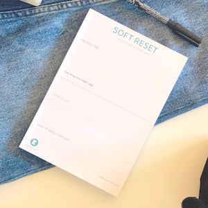 Könnte beinhalten: Ein weißer Papierblock mit der Aufschrift SOFT RESET und Aufforderungen wie Right Now I feel, One thing that might help, What can wait und When I'm ready I'll start with. Der Block liegt auf blauem Jeansstoff neben einem schwarzen Stift.
