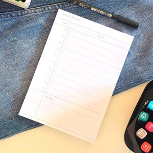 Peut inclure: Un bloc-notes de planning quotidien blanc avec du texte et des lignes gris, comprenant des colonnes pour l'heure de 8 à 17, les tâches et les notes. Le bloc-notes est posé sur du tissu denim bleu avec un stylo noir et des marqueurs.