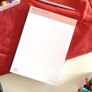 Op de afbeelding: Een wit notitieblok met een perzikkleurige koptekst met de tekst BRAIN DUMP en everything out, nothing organized. Een zilveren pen ligt boven het notitieblok, omringd door kleurrijke stiften in houders op een rode stoffen ondergrond.