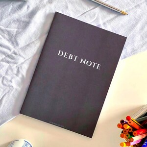 Puede incluir: Un cuaderno gris oscuro con las palabras DEBT NOTE impresas en letras mayúsculas blancas en la portada. El cuaderno está colocado cerca de un bolígrafo plateado, un recipiente blanco lleno de lápices de colores y una botella blanca con tapa azul.