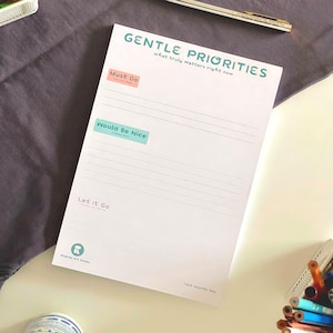 Könnte beinhalten: Ein weißer Papier-Notizblock mit dem Titel Gentle Priorities und den Abschnitten Must Do, Would Be Nice und Let It Go. Der Block enthält türkisfarbenen und pfirsichfarbenen Text, horizontale Linien und den Text rest counts too unten.