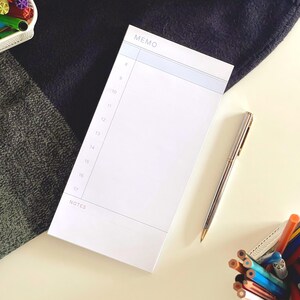 Könnte beinhalten: Ein weißer, rechteckiger Notizblock mit einem hellblauen Kopfbereich und dem Wort MEMO, einer vertikalen Spalte mit Zeitangaben von 8 bis 17 Uhr sowie einem unteren Bereich mit der Aufschrift NOTES. Ein silberner Kugelschreiber liegt daneben.