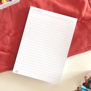 Puede incluir: Un bloc de notas rectangular blanco con líneas horizontales y el texto japonés "メモを取る" impreso en la parte superior. El bloc de notas presenta un pequeño logotipo circular en la esquina inferior izquierda y está rodeado de varios marcadores y lápices de colores.