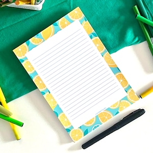 Puede incluir: Un bloc de notas rectangular con un centro blanco y líneas azules horizontales, rodeado por un borde de rodajas de limón amarillas y hojas verdes sobre un fondo azul claro. El bloc está colocado cerca de lápices de colores y un bolígrafo negro.
