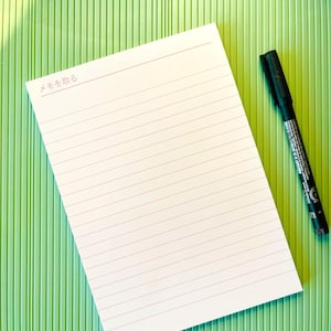 May include: A white notepad with horizontal pink lines and the Japanese text "メモを取る" at the top, placed on a light green scoring board with a visible ruler measuring up to 28 inches. A black felt-tip pen lies to the right of the notepad.