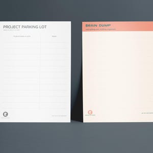 Könnte beinhalten: Zwei Notizblöcke, einer weiß und einer cremefarben, lehnen an einem dunkelgrauen Hintergrund. Auf dem weißen Notizblock steht oben "PROJECT PARKING LOT". Auf dem cremefarbenen Notizblock steht oben "BRAIN DUMP". Beide haben unten ein Logo.