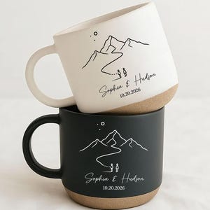 May include: Two ceramic mugs stacked vertically, one white and one black, each featuring a line art mountain landscape, two figures, and the text Sophia & Hudson 10.20.2026. The mugs have a smooth upper finish and a textured, tan-coloured base.