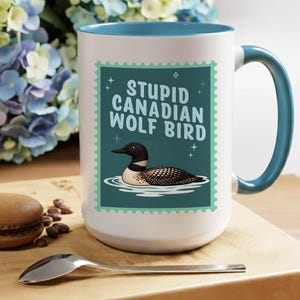 May include: White ceramic mug with a teal interior and handle. The mug features a teal and white stamp design with the text "STUPID CANADIAN WOLF BIRD" and an illustration of a loon. The mug is on a wooden surface.