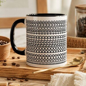 May include: A white ceramic mug with a black interior and handle, featuring a repeating horizontal pattern of black geometric shapes, lines, and semi-circles around the exterior surface of the cylindrical vessel.