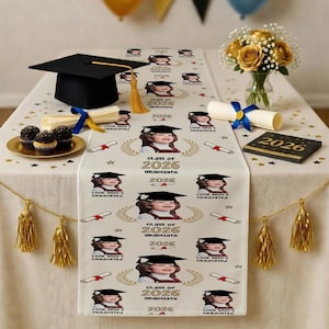 May include: A graduation-themed table setting with a black mortarboard, a table runner printed with a child's photo and the text "Class of 2026 Graduate", cupcakes, and decorative tassels. A bouquet of yellow roses is in a vase.