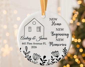 May include: A white ceramic Christmas ornament with a gold ribbon. The ornament features a house illustration, names "Audrey & James", address, and the words "New Home, New Beginning, New Memories". Black floral designs adorn the bottom.