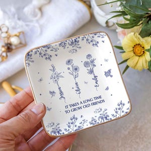 May include: Square ceramic trinket dish with a gold rim, featuring blue floral designs and the names Jessica, Olivia, and Emily. The text "IT TAKES A LONG TIME TO GROW OLD FRIENDS" is also included.