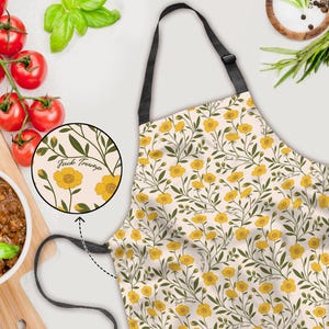May include: Apron with a floral pattern of yellow flowers and green leaves on a cream background. The apron has a black neck strap and waist ties. The text "Fuck Trump" is printed on the apron.