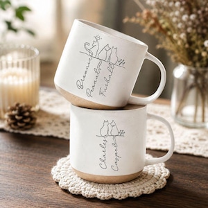 May include: Two stacked ceramic mugs with a speckled white glaze and a natural tan base. The top mug features line art of three cats with the names Brownie, Peanut, and Furball. The bottom mug shows two cats with the names Charles and Casper.
