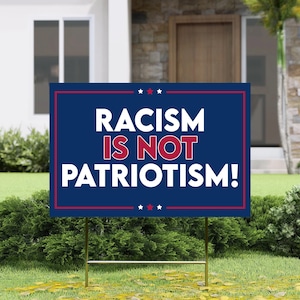 May include: A blue yard sign with the text "RACISM IS NOT PATRIOTISM!" in white and red lettering. The sign has a red and white border and is displayed on a metal stake in a grassy garden.