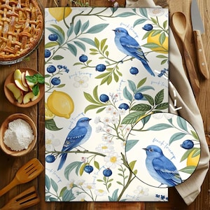 May include: A white kitchen towel with a botanical design featuring bluebirds, lemons, blueberries, and white flowers. The text "fuck Trump" is repeated throughout the design. A pie, apples, and flour are in the background.