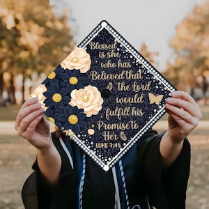 May include: A graduation cap decorated with navy blue and cream-colored flowers with gold centers. The cap has the text "Blessed is she who has Believed that the Lord would fulfill his Promise to Her Luke 1:45."