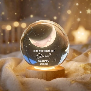 May include: A glass sphere featuring a crescent moon design and the inscription "Beneath This Moon Olivia Was Born 27.08.2026." The sphere is lit from within, resting on a wooden base. The background is blurred with soft, warm lighting.