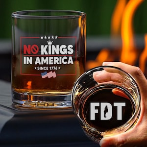 May include: Two clear glass tumblers with a brown liquid inside. The tumbler on the left has the text "No Kings in America *Since 1776*" with a small American flag image. The tumbler on the right has the text "FDT" in white letters.