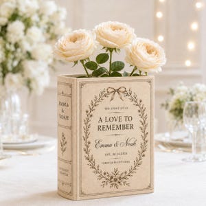 May include: A rectangular beige book-shaped vase featuring a floral wreath design and the text A LOVE TO REMEMBER, Emma & Noah, EST. 06.14.2026, and FOREVER BEGINS HERE. Three cream-colored roses with green leaves are arranged inside the top of the vase.