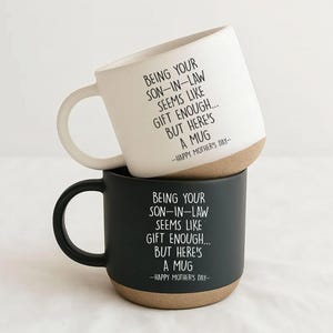 May include: Two ceramic mugs stacked on top of each other. The top mug is white with a brown base and a curved handle. The bottom mug is black with a brown base and a curved handle. Both mugs have the text "BEING YOUR SON-IN-LAW SEEMS LIKE GIFT ENOUGH... BUT HERE'S A MUG -HAPPY MOTHER'S DAY-" printed on them.