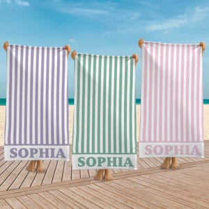 May include: Three beach towels with vertical stripes in purple, green, and pink, each with a white stripe. The towels have the name "SOPHIA" printed in a white rectangular band at the bottom. The towels are held up on a wooden deck with a beach and ocean background.