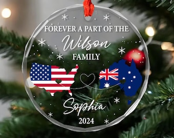 May include: A clear glass Christmas ornament with a red ribbon. The ornament features a map of the United States and Australia connected by a heart and a dotted line. The text "Forever a part of the Wilson Family" and "Sophia 2024" is printed on the ornament.