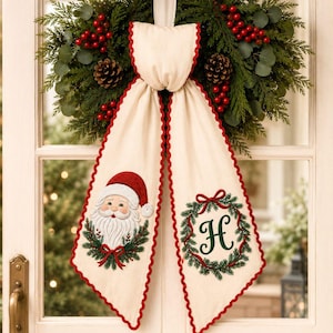 Peut inclure: Une couronne de Noël décorative avec des aiguilles de pin vertes, des pommes de pin et des baies rouges. Un nœud en tissu crème est suspendu au centre, avec une bordure festonnée rouge, un visage de Père Noël brodé et une lettre H verte.