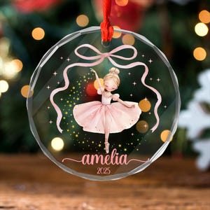 May include: A clear, circular Christmas bauble featuring a ballerina illustration. The ballerina is in a pink tutu, with pink ribbons and stars. The name "amelia" and the year "2025" are written in pink script.