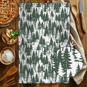 May include: White kitchen towel with a repeating pattern of dark green pine trees. The towel is rectangular and made of a soft fabric. A close-up shows the tree pattern in detail. Kitchen and home decor.