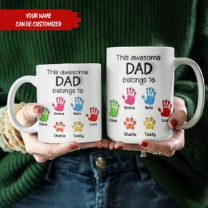 May include: Two white ceramic mugs with the text "This awesome DAD belongs to" and colorful handprints and paw prints. The mugs have the text "YOUR NAME CAN BE CUSTOMIZED" at the top.
