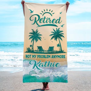 May include: A beach towel with a tropical design featuring palm trees, beach chairs, and the words "Retired, Not My Problem Anymore" and the name "Kathie". The towel is held up against a beach and ocean backdrop.