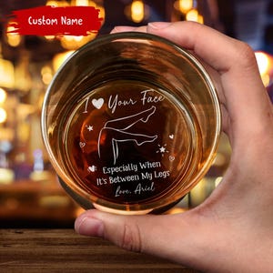 May include: A clear whiskey glass with etched text and a graphic of legs. The text reads "I ♡ Your Face", "Especially When It's Between My Legs", and "Love, Ariel". The glass is held in a hand.