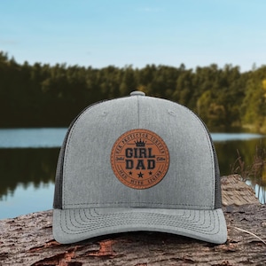 May include: A gray trucker hat with a black mesh back. The front features a circular patch with the words "GIRL DAD" and "HER PROTECTOR FOREVER" in a brown, vintage-style design. The hat is sitting on a wooden surface.