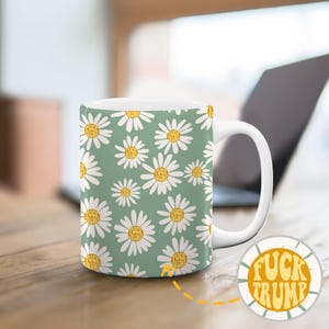 May include: White ceramic mug featuring a daisy pattern with the words "FUCK TRUMP" in the centre of each flower. The daisies are white with yellow centres, set against a sage green background.