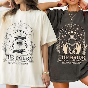 May include: Oversized t-shirts in ivory and pepper colours. Each shirt has a graphic design with hands, a wine glass, and celestial elements. Text on the shirts reads "THE COVEN" and "THE BRIDE", with "Jasmine's Bachelorette, Sedona, Arizona" below.