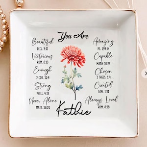 May include: Square white ceramic trinket dish with gold trim. The dish features a red and pink flower illustration and inspirational text including "Beautiful," "Victorious," and "Amazing." The name "Kathie" is also written on the dish.