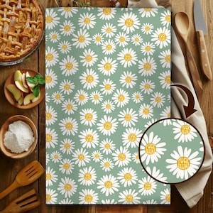 May include: A light green kitchen towel with a daisy pattern. Each daisy has white petals and a yellow center with the words "FUCK TRUMP" in a circular design. The towel is surrounded by kitchen items.