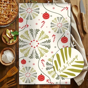 May include: A cream-colored kitchen towel with a festive Christmas design. The towel features green and black stylized tree shapes, red ornaments, candy canes, and snowflakes. The text "Fuck Trump" is incorporated into the design.