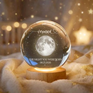 May include: A clear glass sphere with a full moon and starry background, resting on a wooden base. The sphere has the name "Veronica" and the text "THE NIGHT YOU WERE BORN 08.25.2026" engraved on it. The background is softly lit with warm tones.