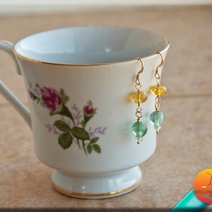 May include: Gold earrings with yellow, green, and blue gemstone beads. The earrings are hanging from a white teacup with a floral pattern.