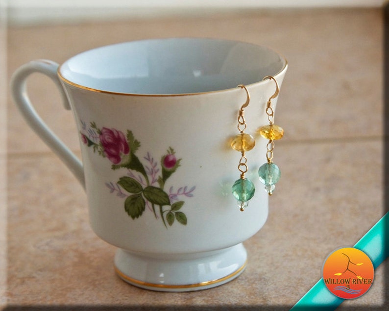 May include: Gold earrings with yellow, green, and blue gemstone beads. The earrings are hanging from a white teacup with a floral pattern.