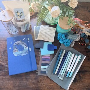 May include: A blue notebook with a dolphin illustration, a glass tumbler with a bamboo lid, a gray pencil case with blue and white pens, a white pouch, a square quote card, sticky notes, washi tape, and a bouquet of white flowers.