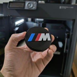 Può includere: Un emblema nero e circolare con il logo BMW in blu, rosso e bianco. L'emblema è tenuto in mano, con una stampante 3D sullo sfondo. Il logo è una "M" stilizzata con i colori BMW.