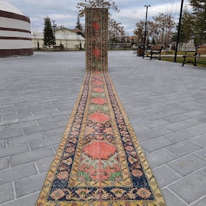 May include: A long, rectangular rug with a traditional design. The rug features a central pattern of red and green floral motifs on a beige background, framed by a dark blue border with intricate patterns. The rug is displayed outdoors on a paved surface.