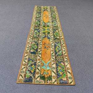 May include: A long, narrow rug with a colorful geometric pattern. The rug has a central orange stripe with blue, green, and white accents. The rug is made of a woven fabric and has a fringed edge.