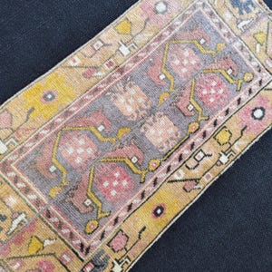 May include: A rectangular, vintage-style rug with a central design in gray, pink, and yellow. The rug features a detailed pattern with geometric and floral elements, framed by a border in shades of yellow and tan. The rug is placed on a dark blue surface.