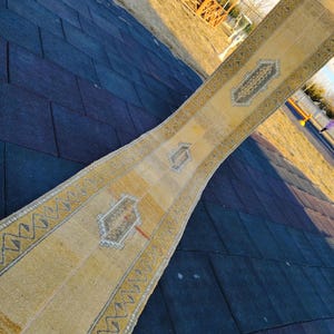 Puede incluir: Una alfombra larga y estrecha de color beige con un patrón geométrico y un medallón central. La alfombra está doblada por la mitad y se encuentra sobre una superficie azul oscuro.